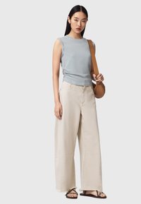 Femme portant un haut sans manches bleu clair, un pantalon large beige, des sandales noires et un sac à bandoulière camel, sur un fond uni.