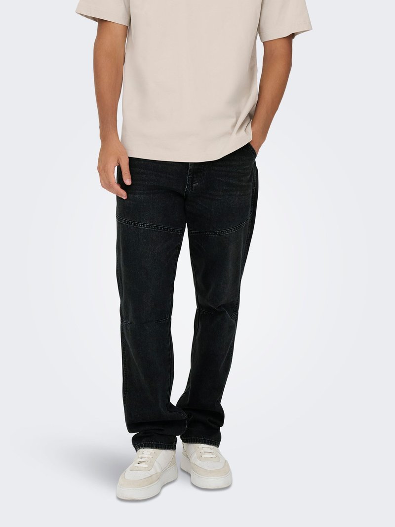 Mann trägt beige kurzärmliges Hemd, schwarze Jeans mit Naht an den Knien und weiße Turnschuhe, steht mit einer Hand in der Tasche.