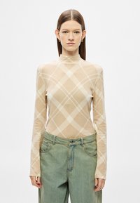 Top bej cu mâneci lungi, cu un model discret în carouri, decolteu tip guler înalt și croială mulată, asortat cu blugi largi, lejeri, de culoare albastru deschis.