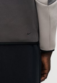 Nike džemperis ar pelēku augšējo daļu un melnu logotipu, kā arī tumšu kontrastējošu apakšējo daļu, izgatavots no teksturētas auduma.