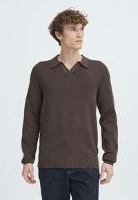 Pulover tricotat maro cu decolteu în V și design cu guler, având mâneci lungi și tiv încrețit, purtat peste blugi închis.