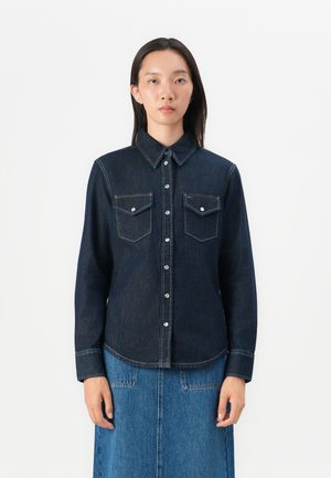 Mørk denimskjorte med lange ermer, knapper foran, to brystlommer og kontraststing. Kombinert med et matchende denimskjørt.