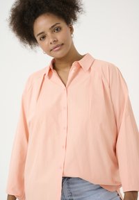 Camisa de gran tamaño en color rosa claro, con botones en el frente, cuello y con hombros caídos; hecha de una tela suave, con una textura sutil y un corte relajado.