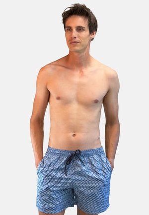 Homme torse nu debout, mains dans les poches, portant un short de bain bleu à motifs avec cordon, de face sur un fond uni.