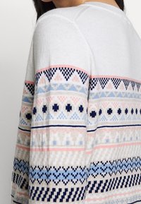 Jersey de punto en gris claro con bandas de patrones multicolores. Presenta diseños geométricos en azul marino, rosa, azul claro y beige a lo largo de las mangas.