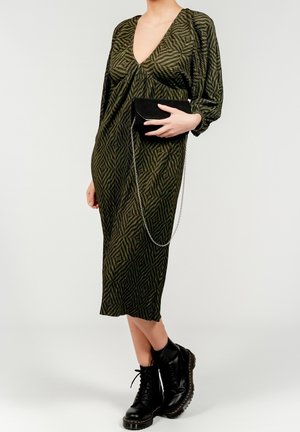 Femme portant une robe midi à motifs verts, des bottes de combat noires, et tenant une pochette noire avec une chaîne argentée sur un fond uni.