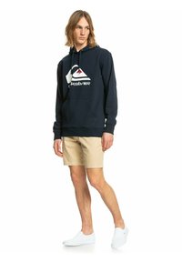 Navy blue hoodie with a large white Quiksilver logo and red accent, made of soft fabric, featuring a kangaroo pocket and drawstring hood.