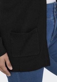 Zwarte gebreide cardigan met een zijzak, geribbelde manchetten en onderrand, gecombineerd met blauwe jeans, met een close-up van de textuur van de stof.