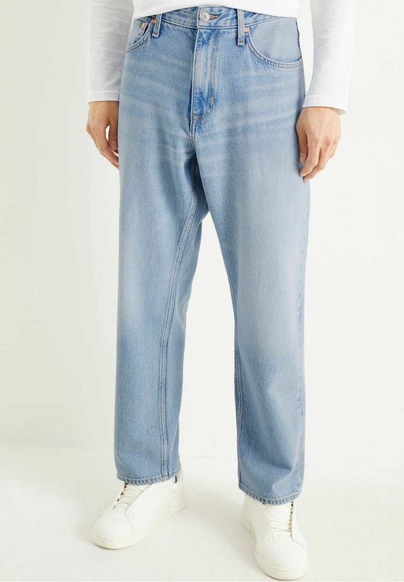 Osoba ubrana w jasnoniebieskie proste jeansy, biały długi rękaw i białe tenisówki, stojąca na jednolitym tle.