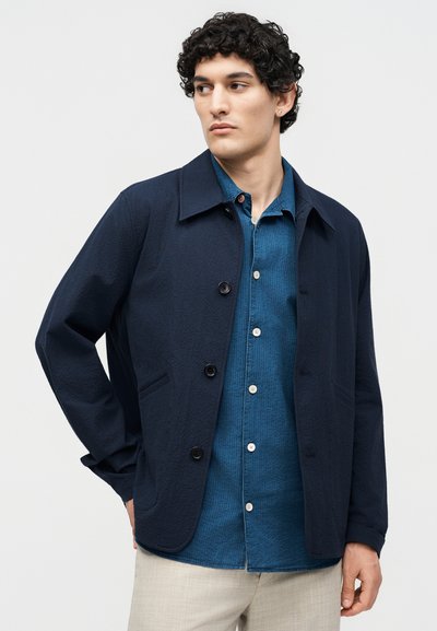 Άνδρας με σγουρά μαλλιά που φοράει σκούρο ναυτικό μπουφάν πάνω από μπλε πουκάμισο με κουμπιά και ανοιχτό μπεζ παντελόνι, κοιτώντας προς το πλάι, σε απλό φόντο.