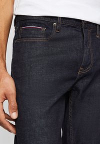 Hombre con jeans azul oscuro con costuras naranja y un pequeño detalle rojo, blanco y azul en el bolsillo delantero, con la mano reposando a un lado.