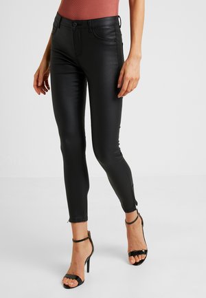 Femme portant un pantalon en cuir noir moulant et des sandales à talons noirs, debout devant un fond clair uni.