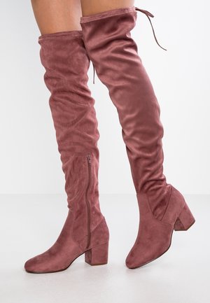 Bottes en suède doux couleur rose poussiéreuse montant au-dessus du genou, avec talons blocs et fermetures éclair latérales, portées sur des jambes contre un fond blanc uni.