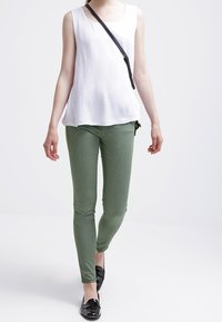 Valkoinen hihaton toppi yhdistettynä oliivinvihreisiin slim-fit-housuihin. Mustat kiiltävät loafers-kengät ja ohut musta olkalaukku viimeistelevät asun.