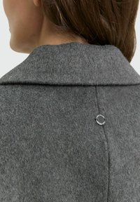 Achteraanzicht van een persoon in een donkergrijze wollen jas met een kleine ronde zwart metalen tag op de bovenste rugnaad.
