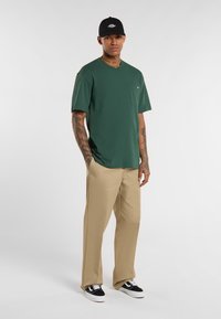 Zelené bavlněné tričko s náprsní kapsou, v kombinaci s béžovými kalhotami rovného střihu a černými teniskami, doplněné o černou čepici.