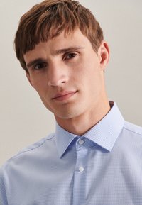 Camicia a quadri azzurro chiaro con colletto classico, taschino sul petto e bottoni bianchi. Il tessuto sembra liscio e fresco.