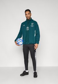 Sinivihreä urheilutakki valkoisin yksityiskohdin ja logolla, mustat kapeat housut sivuraidoilla, pitää moniväristä jalkapalloa kädessään, seisoo yksiväristä taustaa vasten.