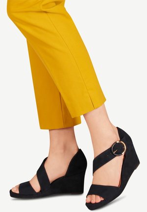 Black suede wedge sandals with a peep toe and crisscross straps, featuring a circular buckle accent. Paired with yellow cropped pants.