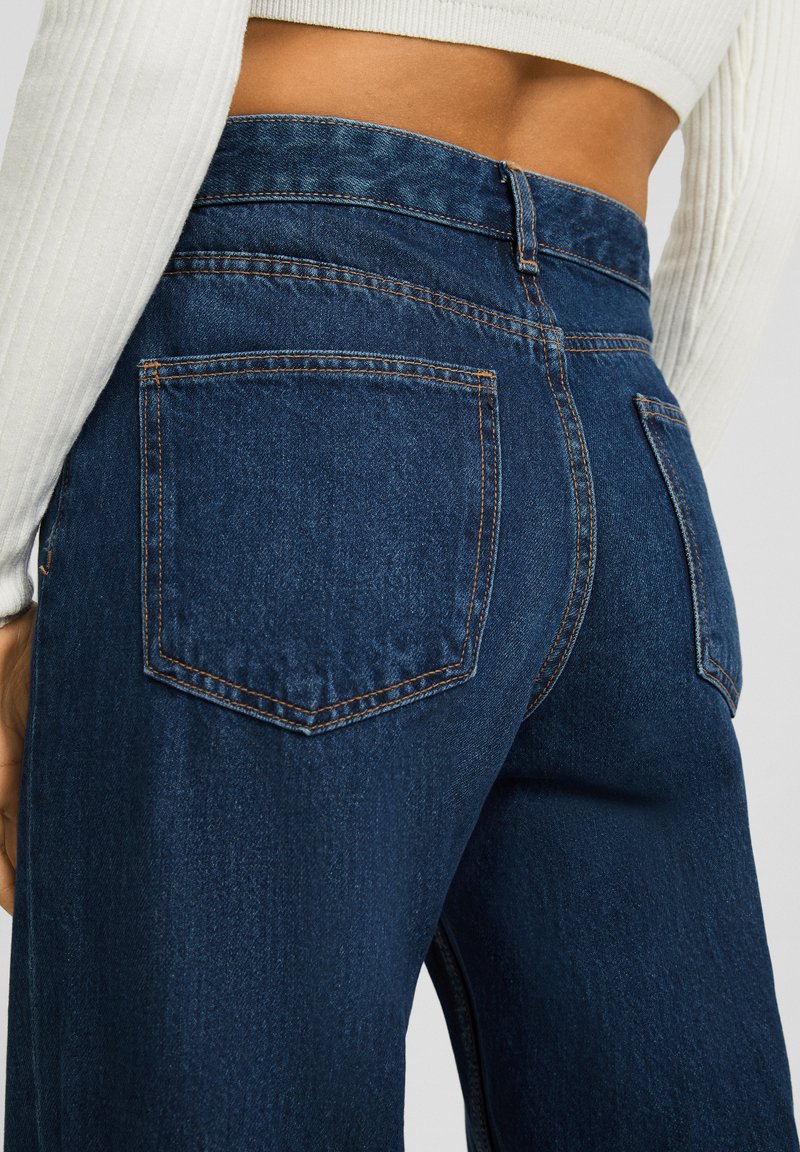 Jeans en denim bleu foncé avec une taille haute, comportant deux poches arrière et des coutures orange contrastantes le long des coutures. Texture lisse.