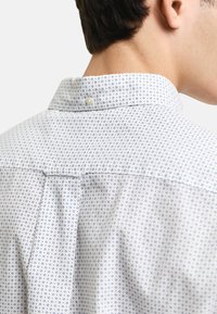 Camisa con cuello abotonado de fondo blanco, con un pequeño patrón geométrico azul, un cuello suave y un diseño de pieza en la espalda.