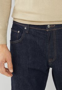 Ciemnoniebieskie dżinsy mają widoczne szwy, przednią kieszeń, metalowy guzik i gładką fakturę, zestawione z beżowym swetrem.