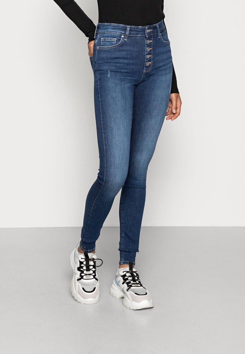 Personne portant un jean skinny bleu taille haute avec braguette boutonnée, un haut noir à manches longues, des baskets blanches épaisses, debout sur un sol gris.