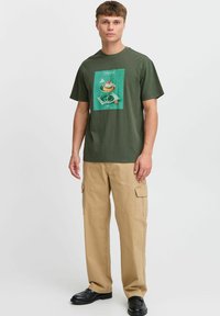 Camiseta de algodón verde oliva con estampado gráfico de un postre en un plato. Combinada con pantalones cargo beige y zapatos negros.