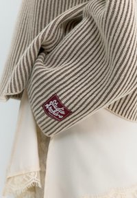 Écharpe en tricot rayée beige et marron avec une étiquette rouge "Studio Arpine Pacific Republic" drapée sur un tissu crème avec bordure en dentelle.