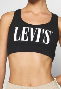 Svart bomullssport-bh med breda band, med en vit "LEVI'S" logotyp tvärs över framsidan. Mjuk textur, åtsittande design, midjehögt snitt.