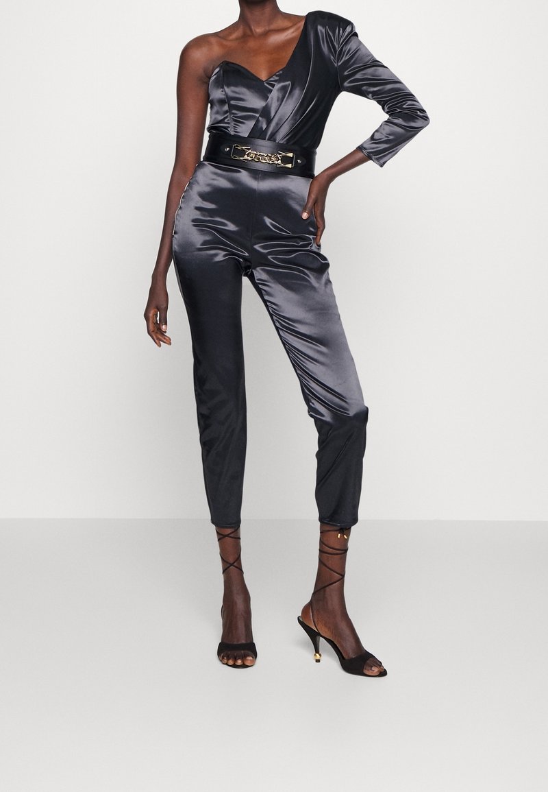 Femme portant une combinaison en satin noire à une épaule avec une large ceinture et des sandales à talons noires à lacets, debout devant un fond uni.