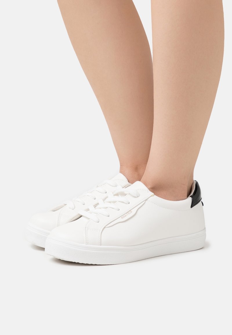 Paire de baskets basses blanches avec lacets blancs et languette de talon noire portées pieds nus sur un fond uni.