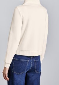 Sudadera color crema con cuello alto, tela suave y puños acanalados. Combinada con vaqueros oscuros de talle alto, mostrando detalles traseros.