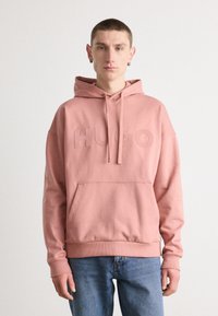 Rosa Kapuzenpullover mit einer Vordertasche und geprägtem "HUGO"-Logo. Mit gerippten Bündchen und Saum, kombiniert mit blauen Jeans.