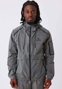 Giacca impermeabile grigia con zip frontale, cappuccio e polsini elasticizzati. Il design include dettagli neri e tasche laterali per la conservazione.