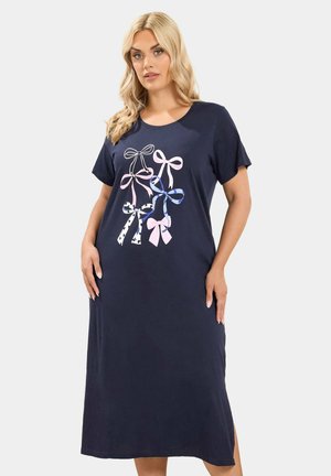 Blonde Frau trägt ein marineblaues Midi-Kleid mit kurzen Ärmeln und rosa, blauen sowie weißen Schleifenmotiven auf der Vorderseite.