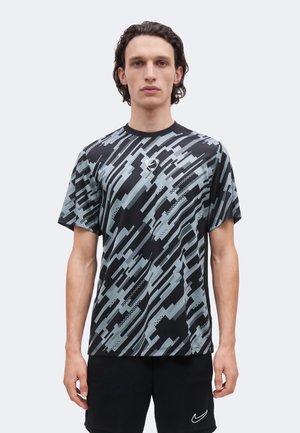 Homme aux cheveux bruns, debout face à nous, portant un t-shirt noir et gris à motif abstrait et un short noir avec un petit logo blanc.