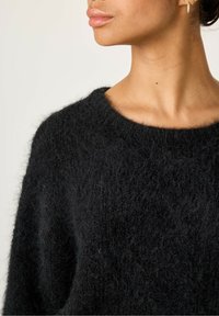 Juodas megztinis su minkšta, pūkuota tekstūra ir plačiu iškirpimu, pasižymintis laisvu siluetu ir matomomis siuvimo detalėmis.