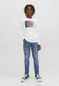 Balta garām piedurknēm krekls ar bloku grafiku, kurā rakstīts "JACK & JONES" sarkanā un jūras zila krāsā, apvienots ar zilām biksēm un jūras zila krāsas sporta apaviem.