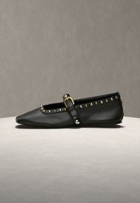 Chaussure plate en cuir noir à bout pointu, bride à boucle dorée et petits clous argentés et dorés le long des bords, exposée sur une surface neutre.