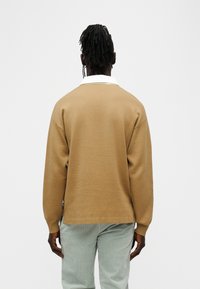 Καφετί πλεκτό πουλόβερ με λευκό γιακά, σε άνετη εφαρμογή και μακριά μανίκια. Το ύφασμα φαίνεται λείο με διακριτή υφή. Φορεμένο με ανοιχτοπράσινο παντελόνι.