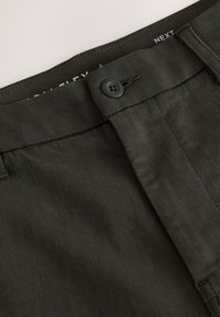 Gros plan sur la ceinture et le bouton d’un pantalon gris foncé avec une couture visible et une partie de l’étiquette intérieure de la marque indiquant "NEXT."
