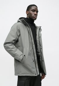 Sivý páperový kabát s kapucňou, obsahujúci zips a uzávery na cvoky, bočné vrecká a hladký, štruktúrovaný povrch.