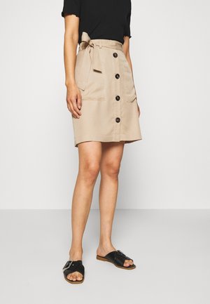 Mujer con falda beige abotonada por delante con lazo lateral, top negro y sandalias negras tipo slide con hebillas, de pie sobre suelo gris.