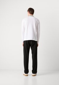 Hvit langermet skjorte med en liten utskjæring bak, kombinert med svarte bukser og lyse sneakers. Enkelt, minimalistisk design.