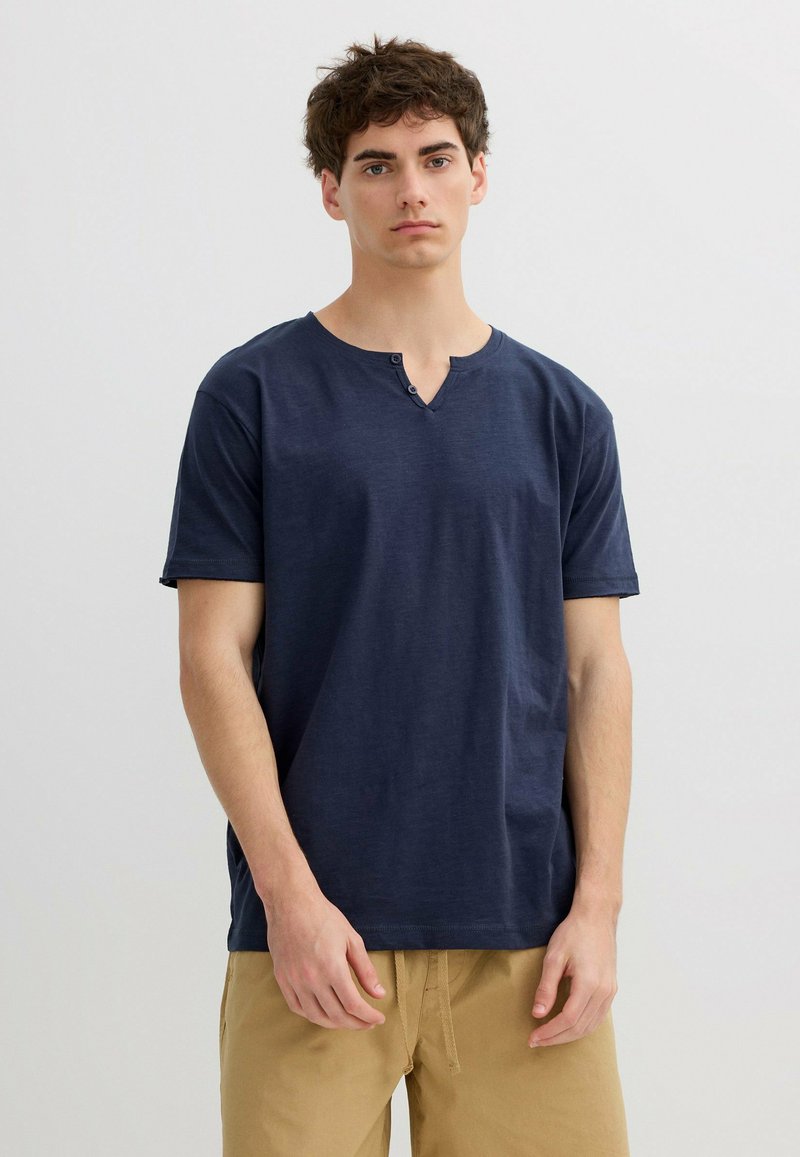 Joven con cabello corto y rizado, vistiendo una camisa Henley de manga corta azul marino y pantalones cortos marrón claro, de pie frente a un fondo liso.