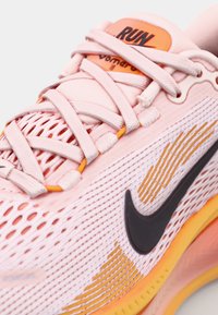Las zapatillas de running Nike en color rosa presentan una parte superior de malla transpirable, un swoosh negro, acentos naranjas y una suela texturizada con detalles amarillos.