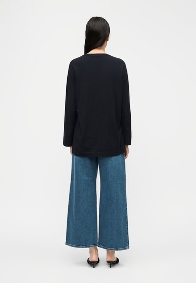 Femme aux longs cheveux bruns portant un chemisier noir à manches longues, un jean large bleu et des chaussures noires à talons, debout de dos.