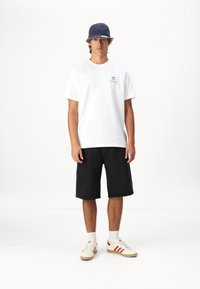 Hvit bomulls t-skjorte med blå grafikk, svarte denimshorts, røde og kremfargede stripete joggesko, marineblå caps. Modellen står mot en ensfarget bakgrunn.