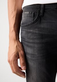 Jeans negre din denim cu o finisare ușor decolorată, având un buzunar frontal și un etichetă de marcă pe spate. Mână odihnindu-se pe coapsă.
